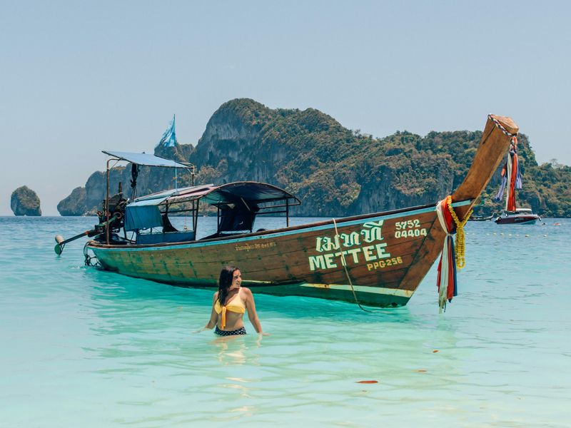 Cette image montre une chaloupe traditionnelle thaïlandaise en bois, avec des falaises de calcaire en arrière-plan. À l'avant, une jeune fille aux cheveux noirs se tient dans l'eau avec un haut de bikini jaune. Les îles Phi Phi sont un must à ajouter à votre itinéraire de 5 jours à Phuket.