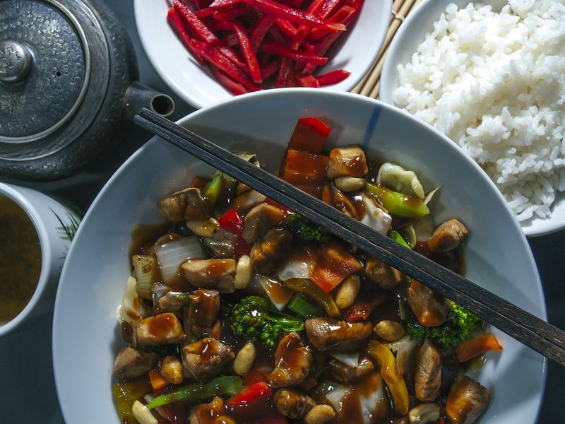 Le poulet Kung Pao est un plat clé de la culture alimentaire chinoise. Ici, le poulet est coupé en morceaux de taille moyenne et mélangé à des légumes dans une sauce brune épaisse. Une paire de baguettes est posée horizontalement au-dessus du bol.