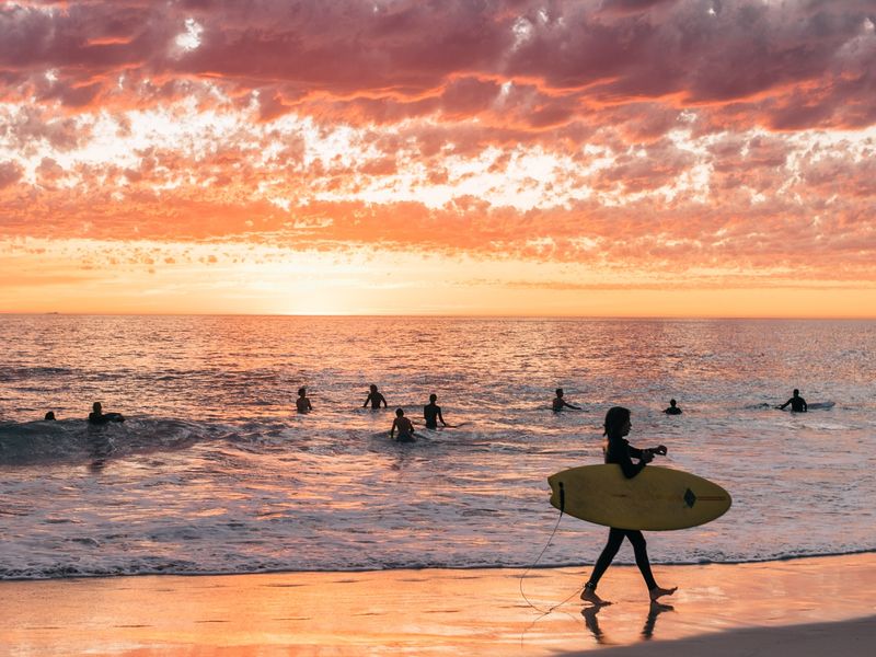 Un surfeur marche avec sa planche de surf sous un bras tandis que d'autres surfeurs attendent une vague. Un surf au lever du soleil mérite une place dans votre itinéraire d'une journée à Nusa Lembongan.