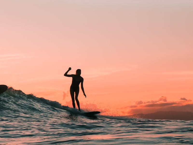 La silhouette d'une femme attrapant une vague sur une planche de surf dans un ciel couleur pêche.