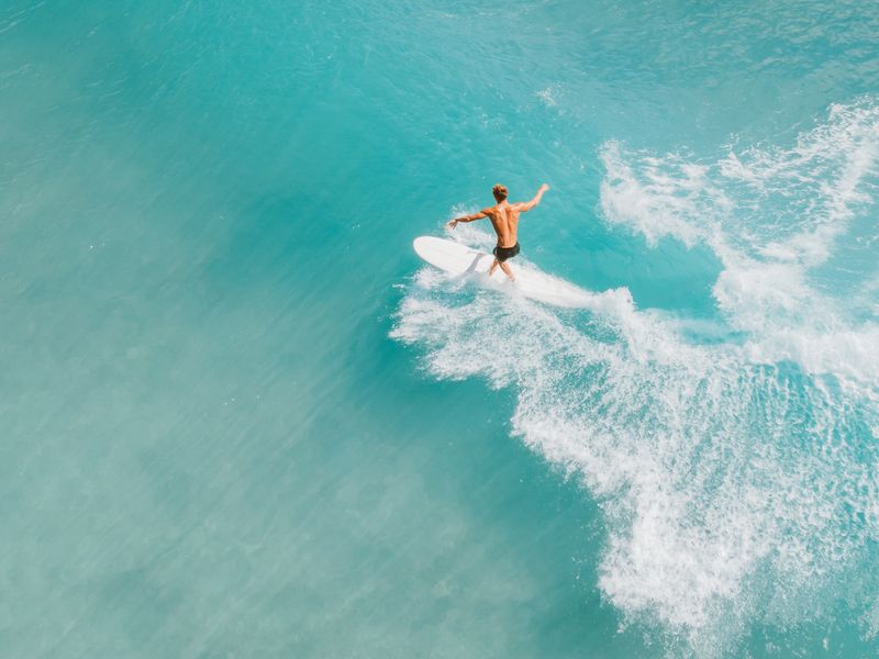 Une photo aérienne d'un homme seul en train de surfer dans des eaux bleues claires. Le Mexique possède certaines des meilleures plages pour le surf au Mexique pour les débutants.