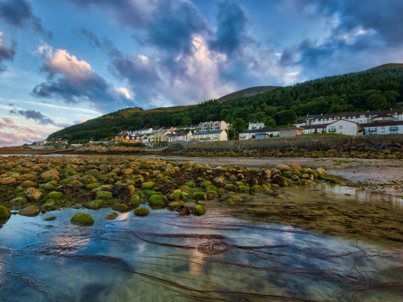 Le plus souvent, vous commencerez votre randonnée du Slieve Donard à partir de Newcastle. Cette image montre des maisons de bord de mer avec en toile de fond une forêt et des sommets de montagne.