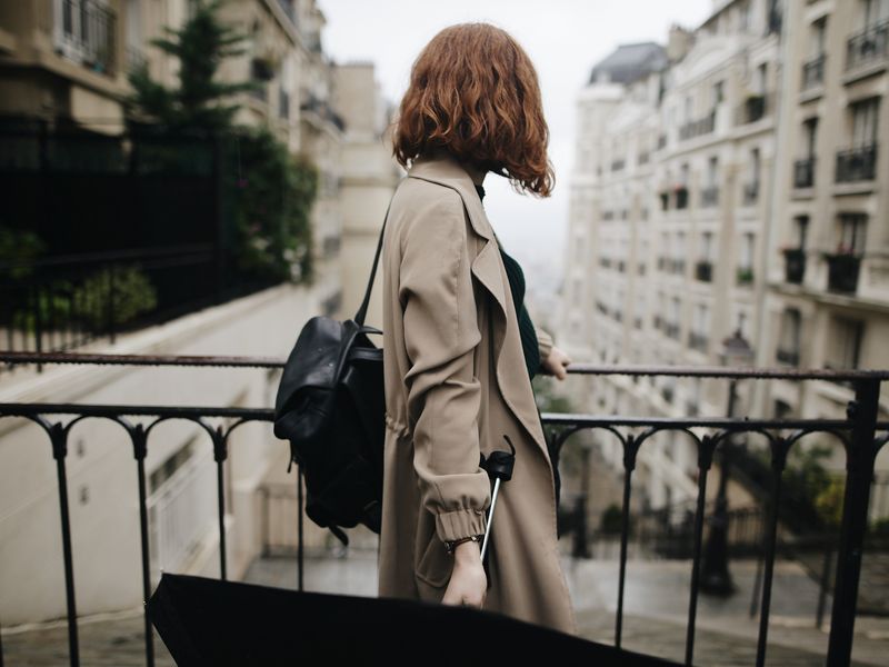 Le trench-coat est un élément historique de la culture vestimentaire britannique. Ici, une femme aux cheveux bouclés regarde par-dessus une balustrade dans un trench-coat beige en tenant un parapluie noir. 
