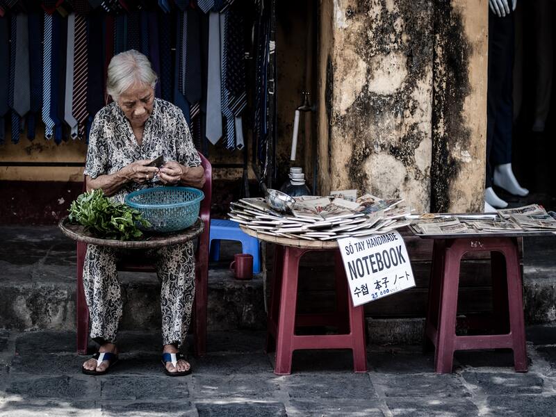 Femme assise sur une chaise, tranchant des légumes tout en vendant des cahiers.